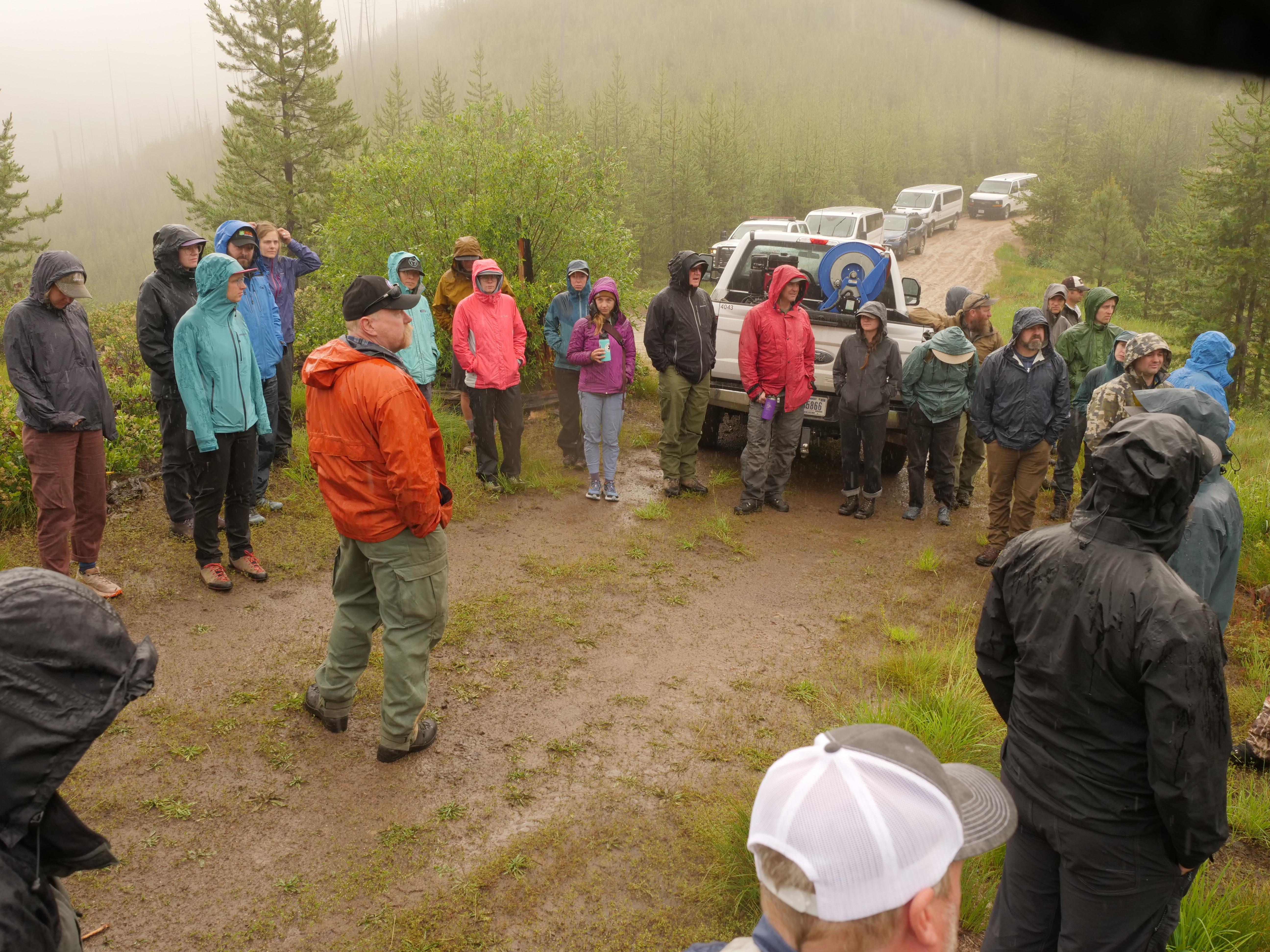 People in rain jackets and hoods standing in a circle on a dirt road in a coniferous forest